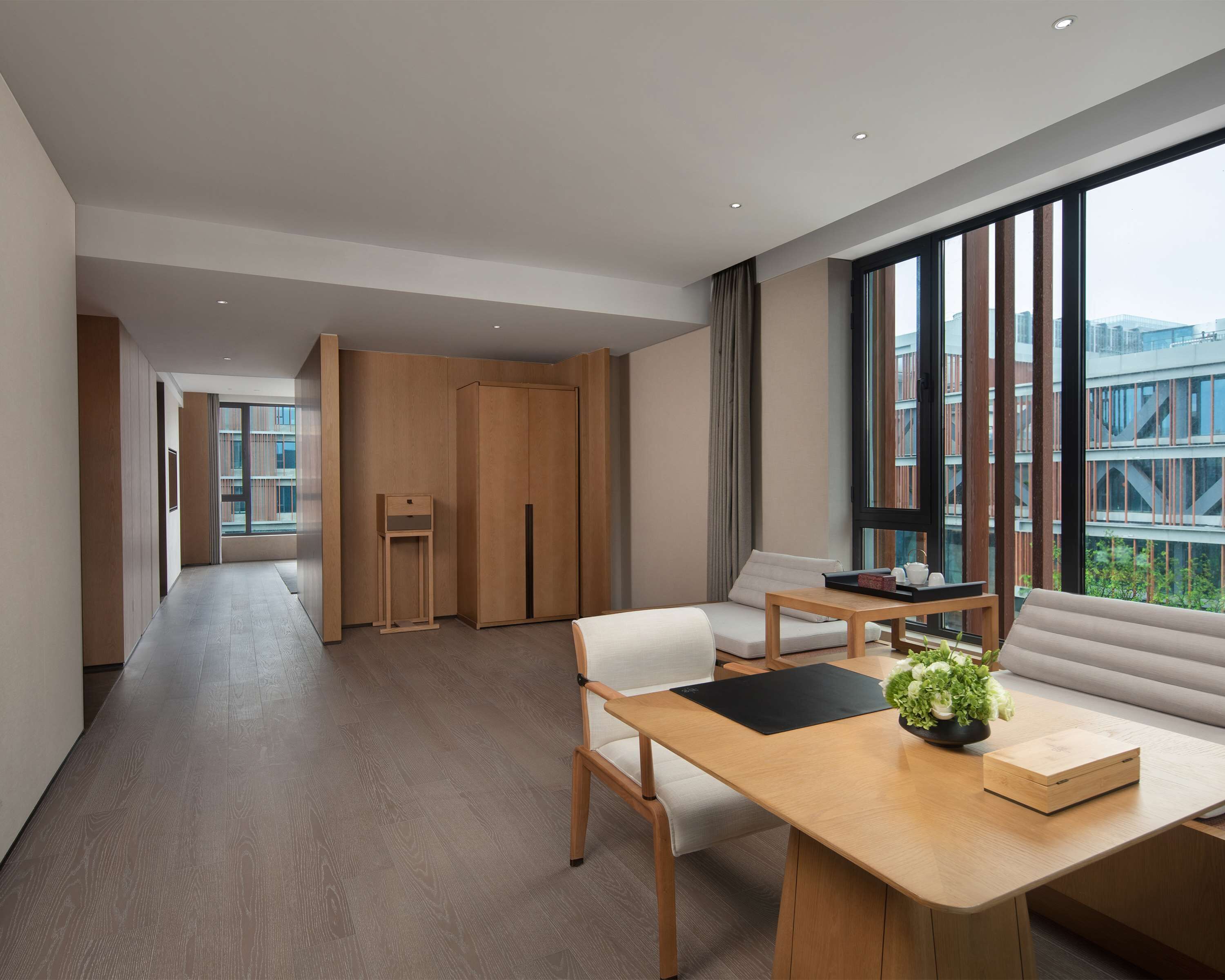 Una habitación de hotel moderna con suelo de madera, un ventanal con vistas a la ciudad, un escritorio de madera con una planta y un sofá.