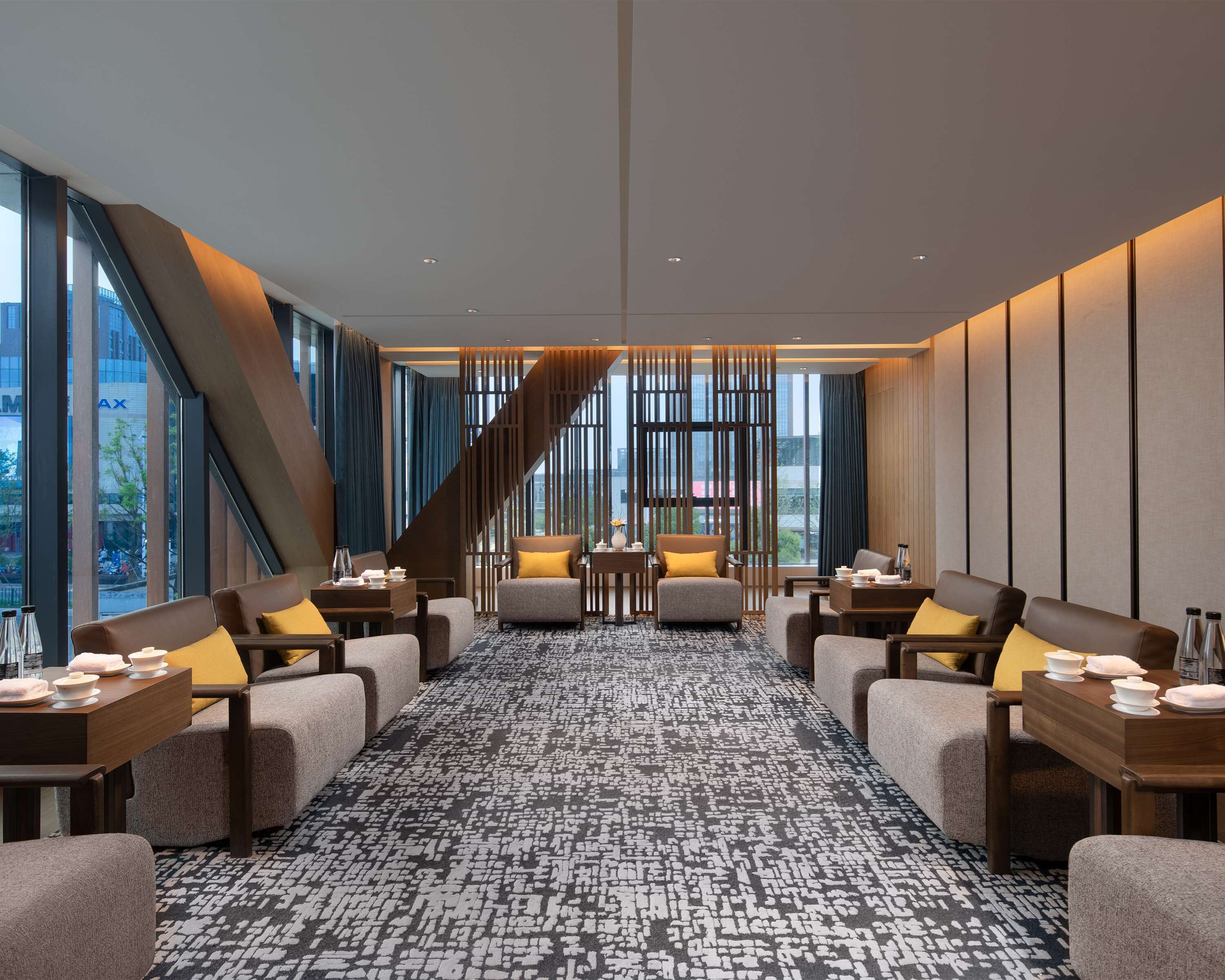 A modern hotel lobby with a central staircase, comfortable seating areas, and large windows offering city views.