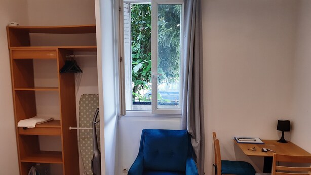 Ein Schlafzimmer mit einem Bett, einem blauen Sessel, einem Holztisch mit Lampe, einem Fenster mit Vorhängen und einem Kleiderschrank.
