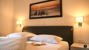 A hotel room with a bed, two bedside lamps, a framed picture on the wall, and a nightstand with a lamp. - Refugium 2 (Kellenhusen)