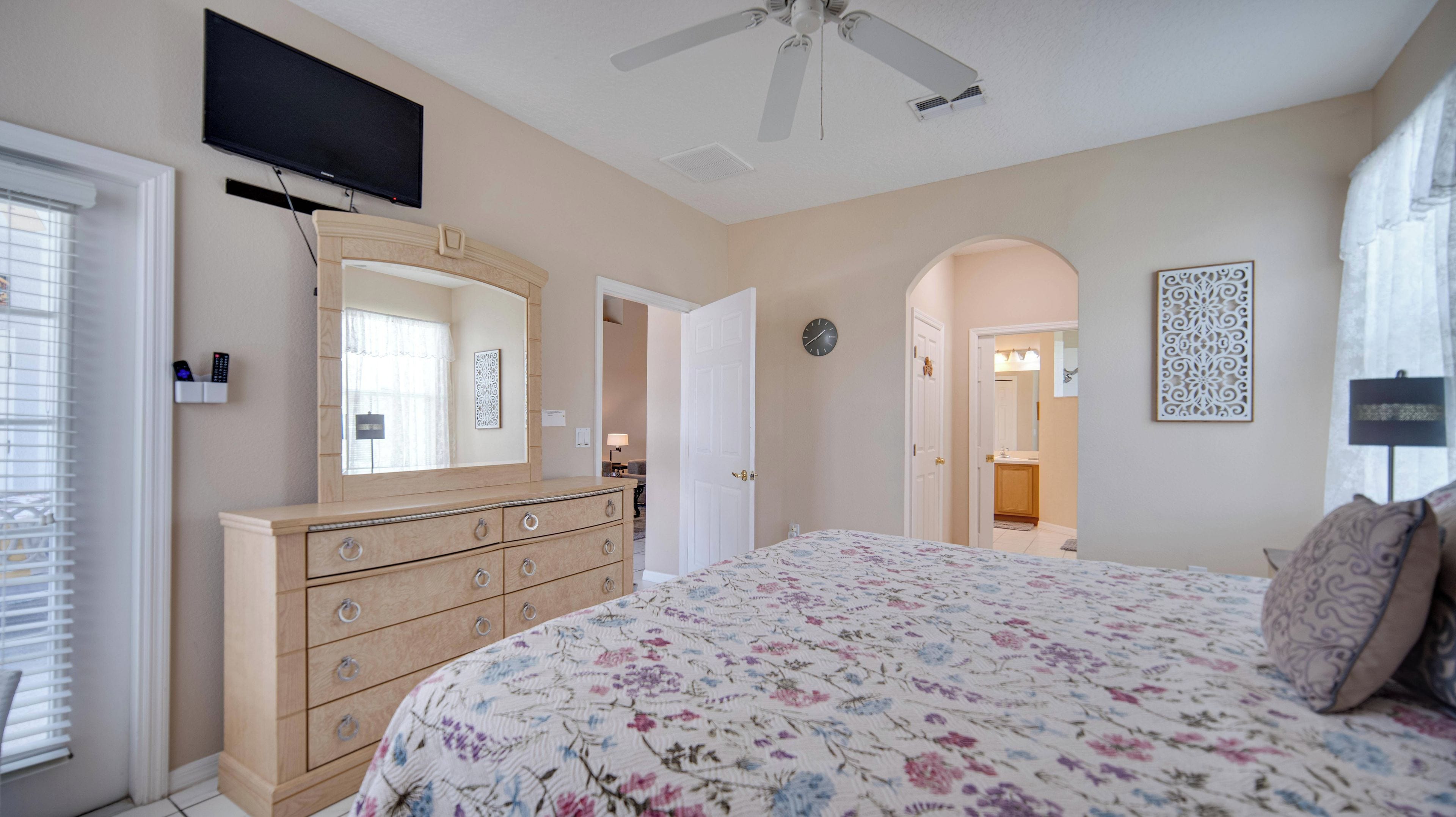 Une chambre à coucher avec un lit, une commode, un ventilateur de plafond, une télévision, et une salle de bain visible à travers une porte ouverte.