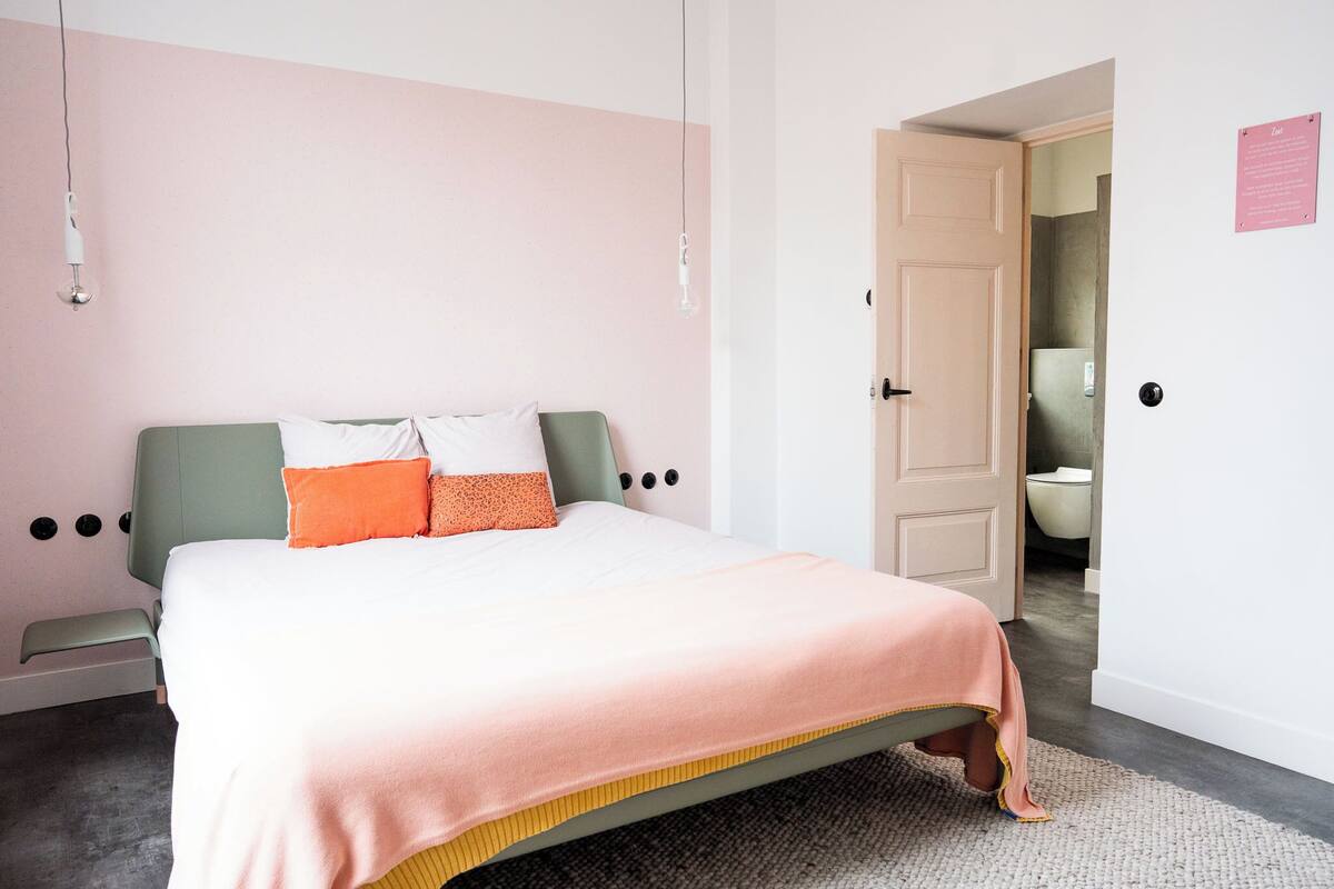 Un dormitorio con cama, un banco, una puerta y un baño visible a través de una puerta abierta.