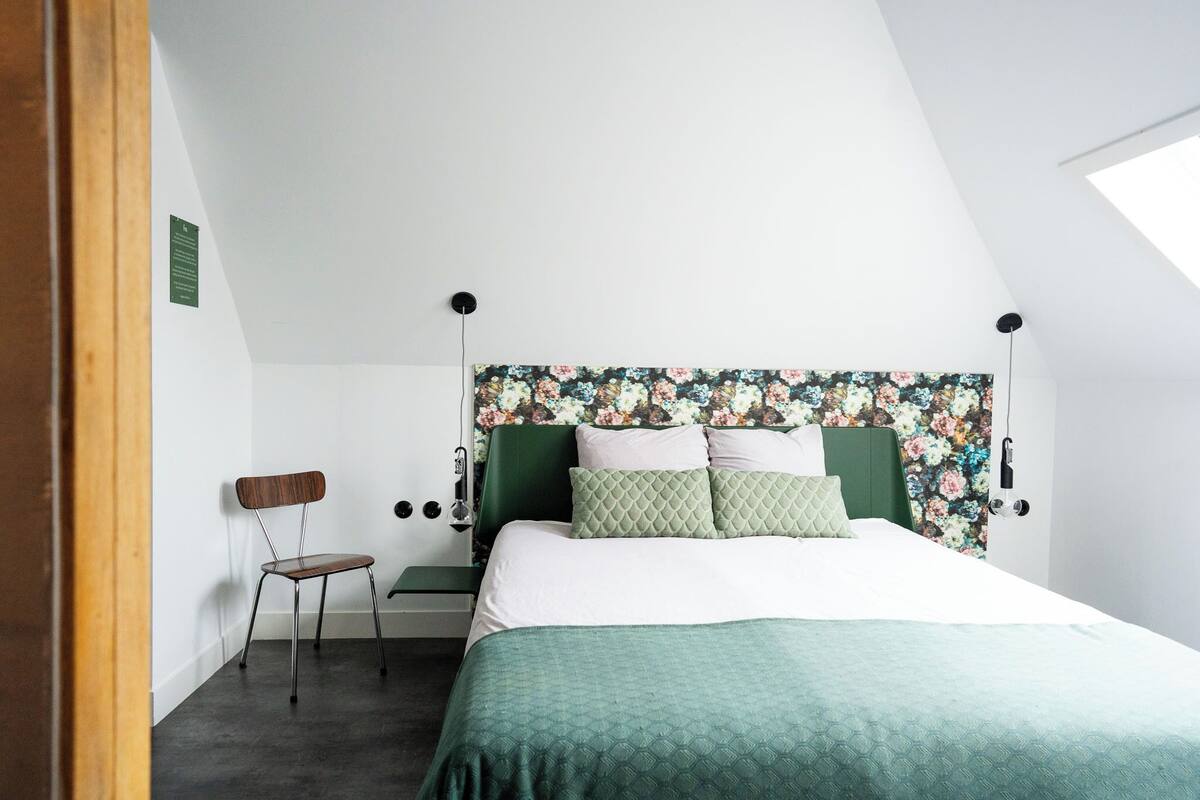 Un dormitorio con colcha verde, cabecero floral, una silla de madera y una mesita de noche pequeña.