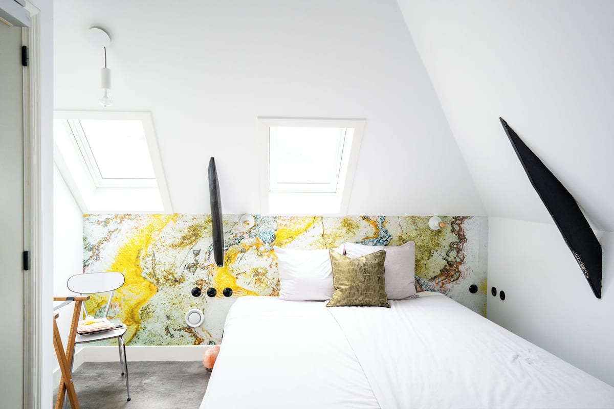 Un dormitorio con techo inclinado, dos tragaluces, una cama con ropa blanca, una silla y una pared con un diseño abstracto y colorido.