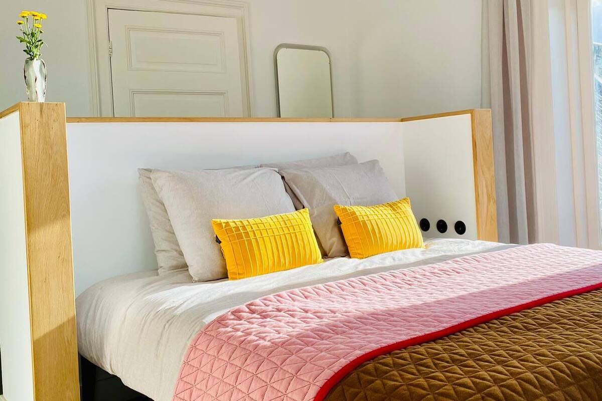 Un dormitorio con una cama, una mesita de noche con un florero y una ventana con cortinas.