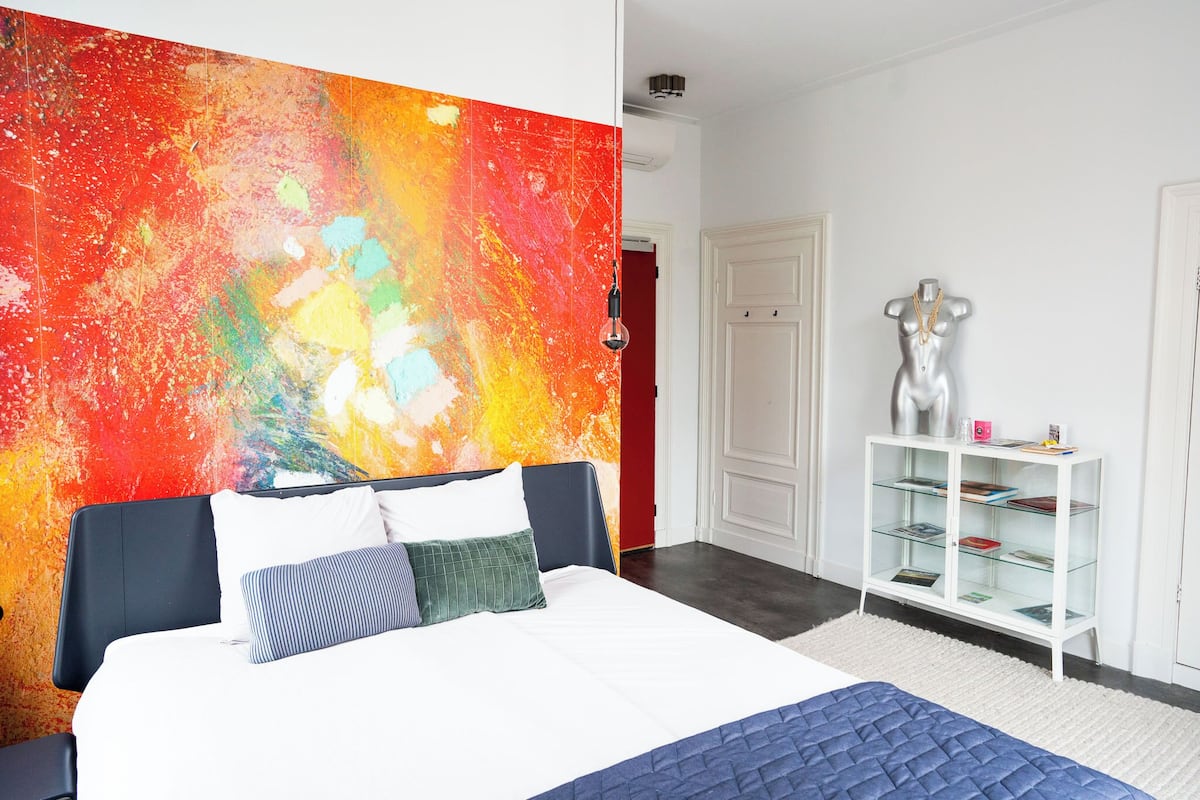 Un dormitorio moderno con una cama grande, un muro decorativo abstracto y lleno de color, un armario blanco y una vitrina de vidrio.