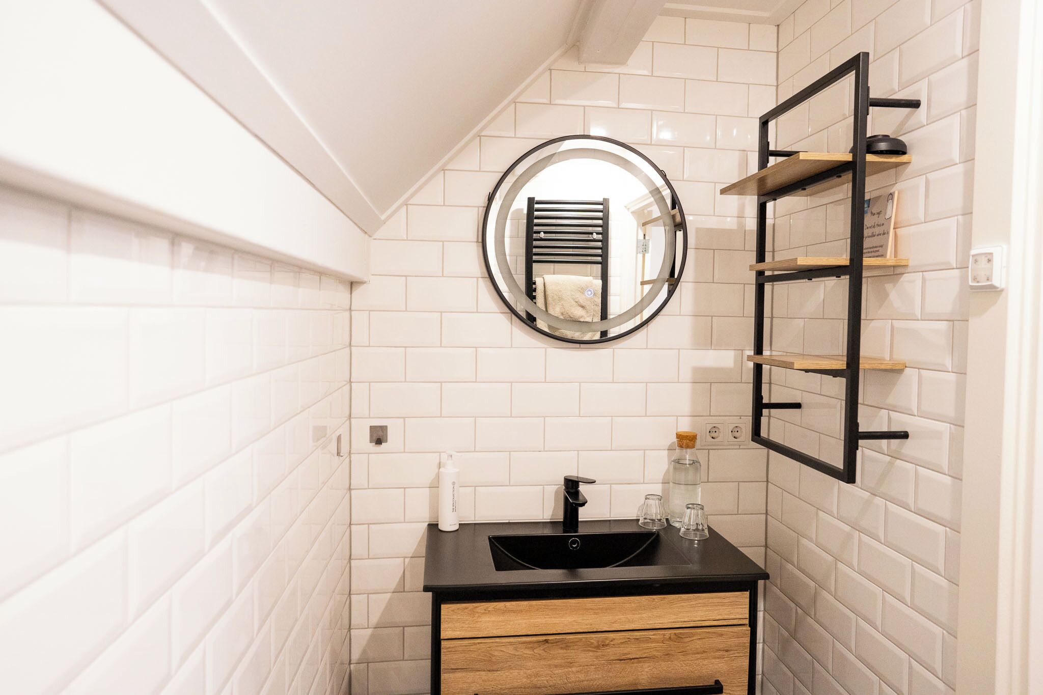 Une salle de bain avec des murs recouverts de carreaux blancs, un lavabo noir, un meuble-lavabo en bois, un miroir rond et une étagère en métal noir.