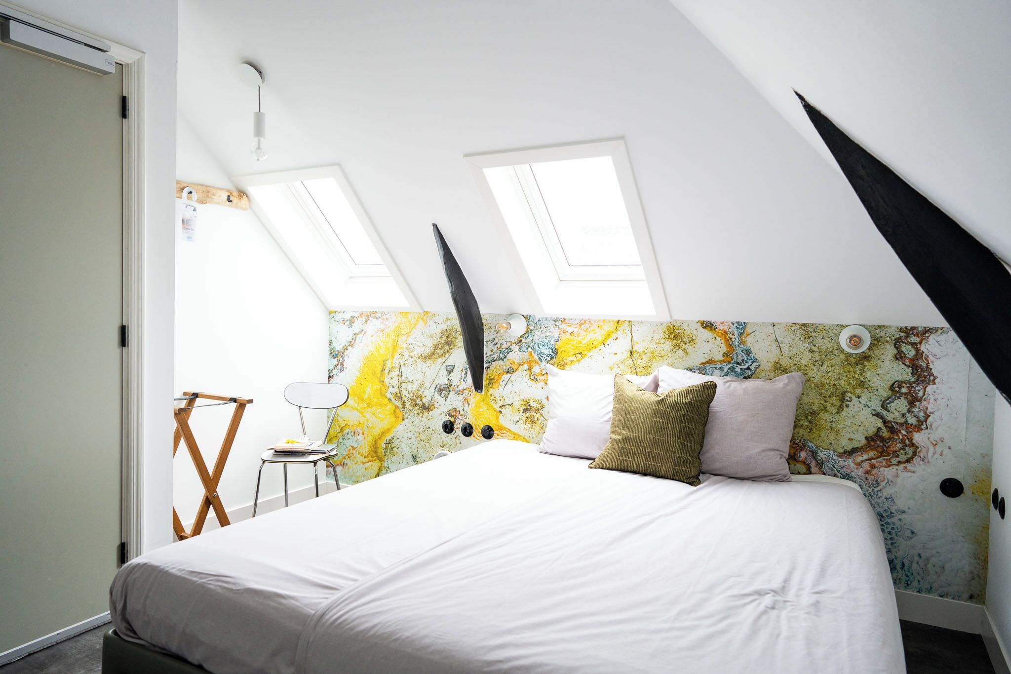 Un dormitorio con techo inclinado, tragaluces, una cama con ropa blanca, una silla y una pared con un diseño abstracto y colorido.