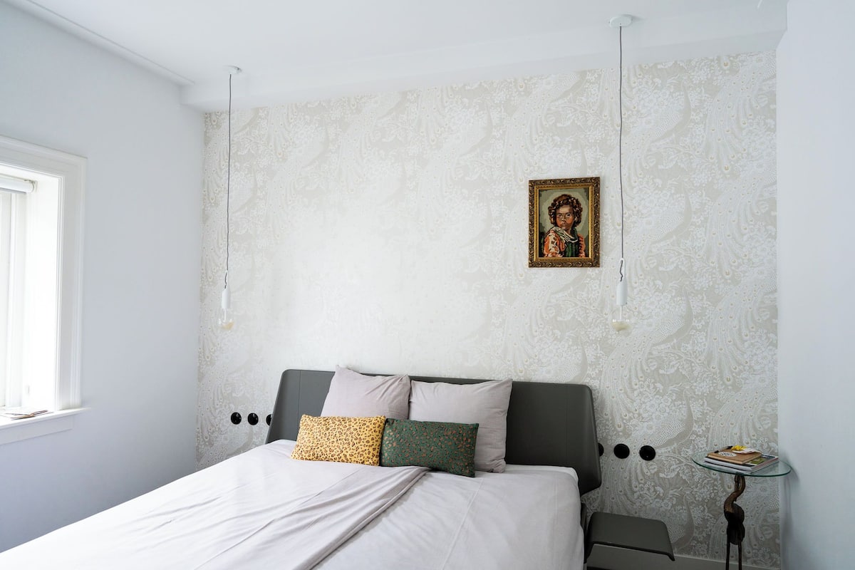 Un dormitorio con cama, almohadas, una mesita de noche, una pequeña mesa y una pared con un diseño decorativo.