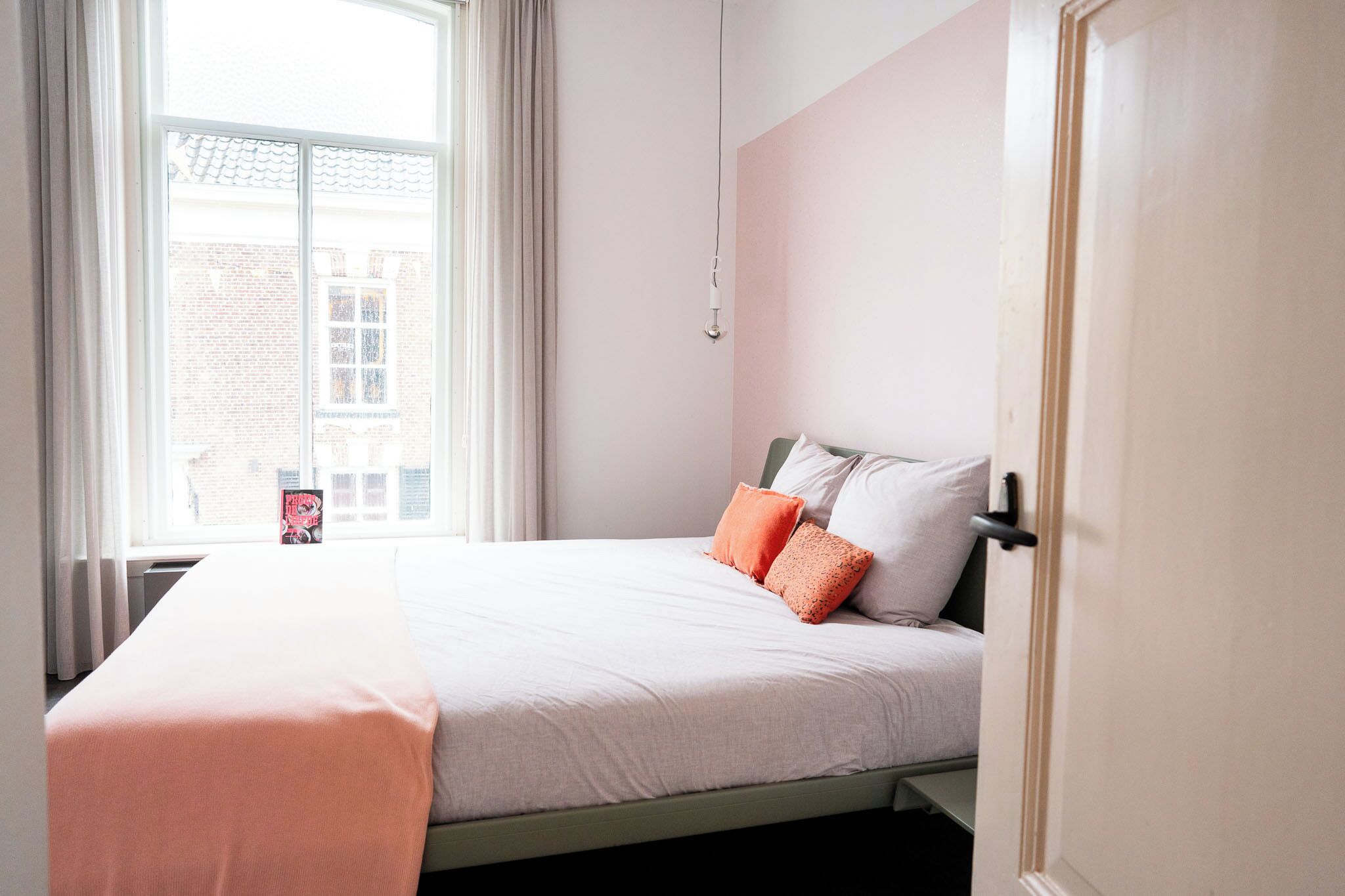 Una cama bien tendida con una manta rosa y dos almohadas, una ventana con cortinas y una puerta a la derecha.