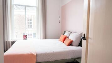 Una cama bien tendida con una manta rosa y dos almohadas, una ventana con cortinas y una puerta a la derecha.