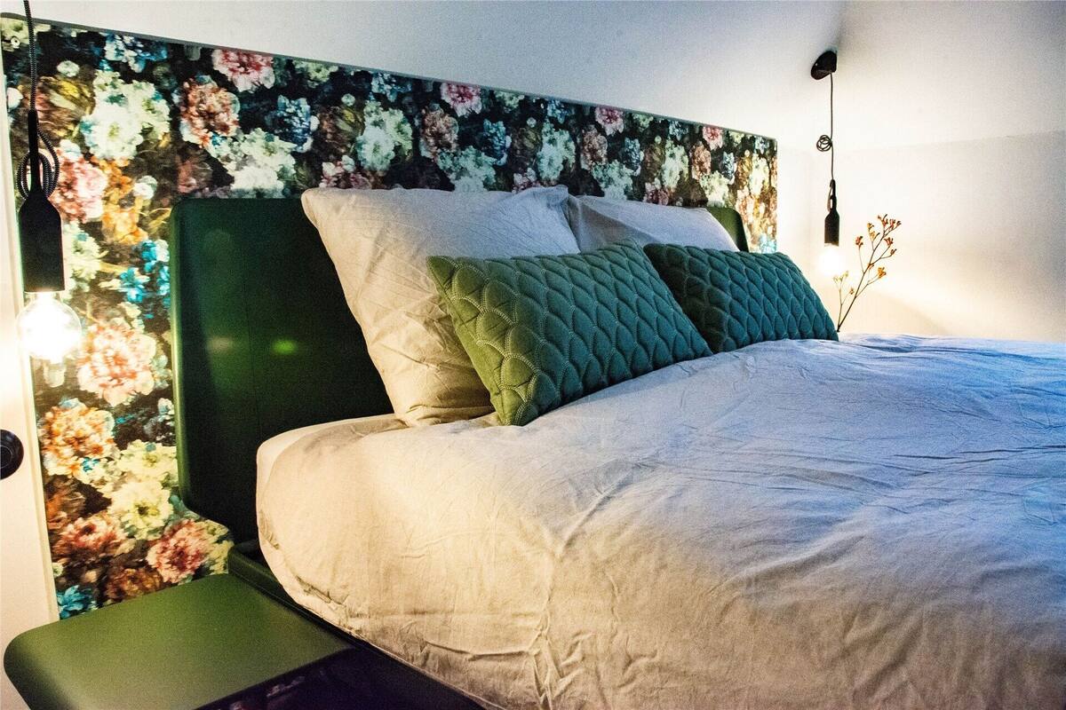 Una cama con cabecera floral, una mesita de noche y una lámpara fijada a la pared.