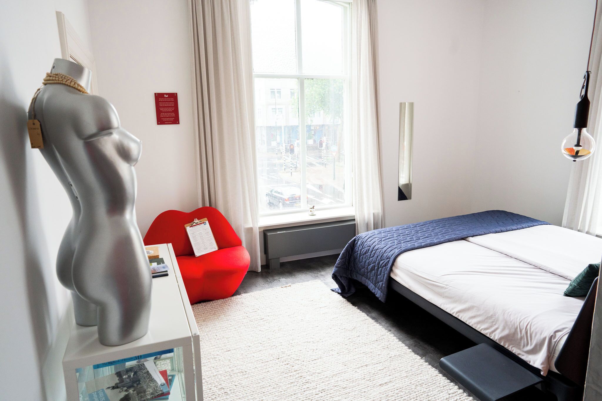Une chambre à coucher moderne avec un lit, un fauteuil rouge, un mannequin et une fenêtre donnant sur la rue.