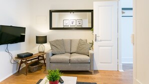 Una sala de estar con un sofá gris, un televisor de pantalla plana, una lámpara y un cuadro enmarcado sobre el sofá.
