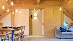 Un espace repas comprenant une table et des chaises en bois, un canapé, et un plafond avec des poutres apparentes et des luminaires suspendus.