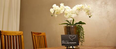 A dining table with a vase of white orchids, a sign with "Willkommen" and a heart symbol, and a modern circular light fixture.