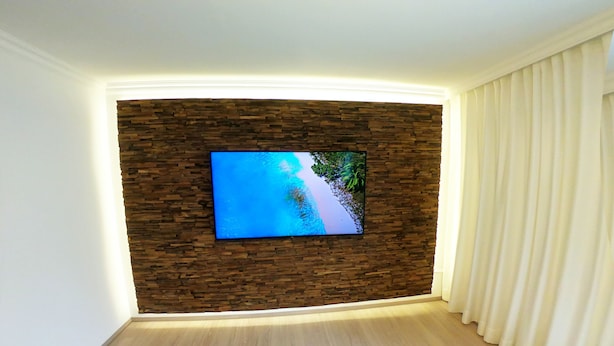 Habitació amb televisió de pantalla plana penjada a una paret de pedra, cortina blanca i una catifes de color clar.