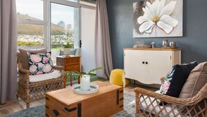 A living room with wicker furniture, a white cabinet, and a large window with a view of a city and hills. - Mouille Point Studio with Breathtaking Views (Cape Town)