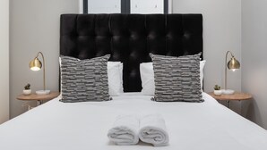 A neatly made bed with white linens, two black and white striped pillows, and a black tufted headboard. There are two bedside tables with lamps and small potted plants. - Edgy Green Point Pad , Balcony , Parking , Pool (Cape Town)