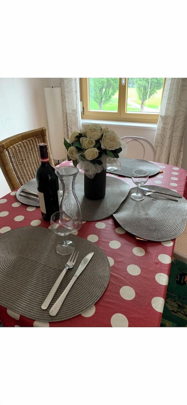 Ein Esstisch, gedeckt mit Platzsets, Besteck, einer Weinflasche, einem Glas und einer Vase mit weißen Blumen.
