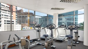 A gym with various exercise machines, large windows, and a view of a cityscape with buildings and construction.