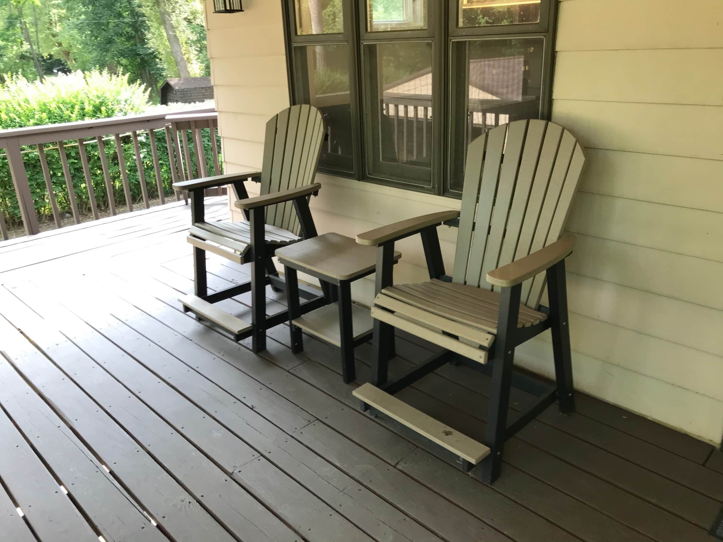 Dos sillas Adirondack en una terraza de madera con barandilla, una ventana con vista a una casa y una cerca de madera al fondo.