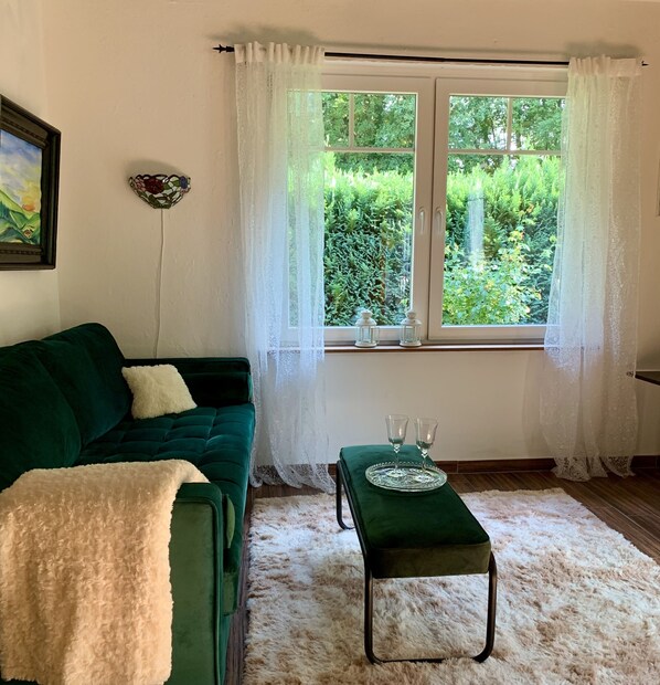 Ein Zimmer mit einem grünen Sofa, einem kleinen Tisch mit zwei Gläsern und einem Fenster mit weißen Vorhängen.