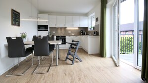 A modern kitchen with white cabinets, a dining area with a table and chairs, and a sliding glass door to a balcony. - Refugium 3 (Kellenhusen)