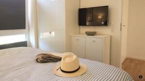 A bedroom with a bed, a television mounted on the wall, and a window with blinds. - Refugium 3 (Kellenhusen)
