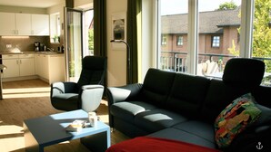 A modern living room with a sofa, armchair, and coffee table. A kitchen is visible in the background with cabinets and a sink. - Refugium 3 (Kellenhusen)