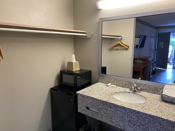 A compact bathroom with a granite countertop, a sink, a mirror, a microwave, and a small refrigerator. - Cabarrus Inn (Concord)
