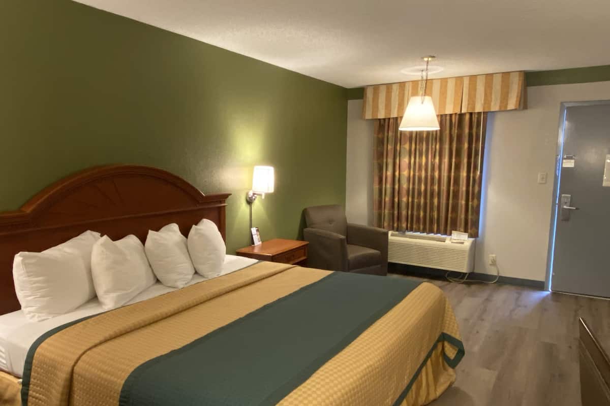 Ett hotellrum med en stor säng, en stol, ett skrivbord, en lampa och ett fönster med gardiner.