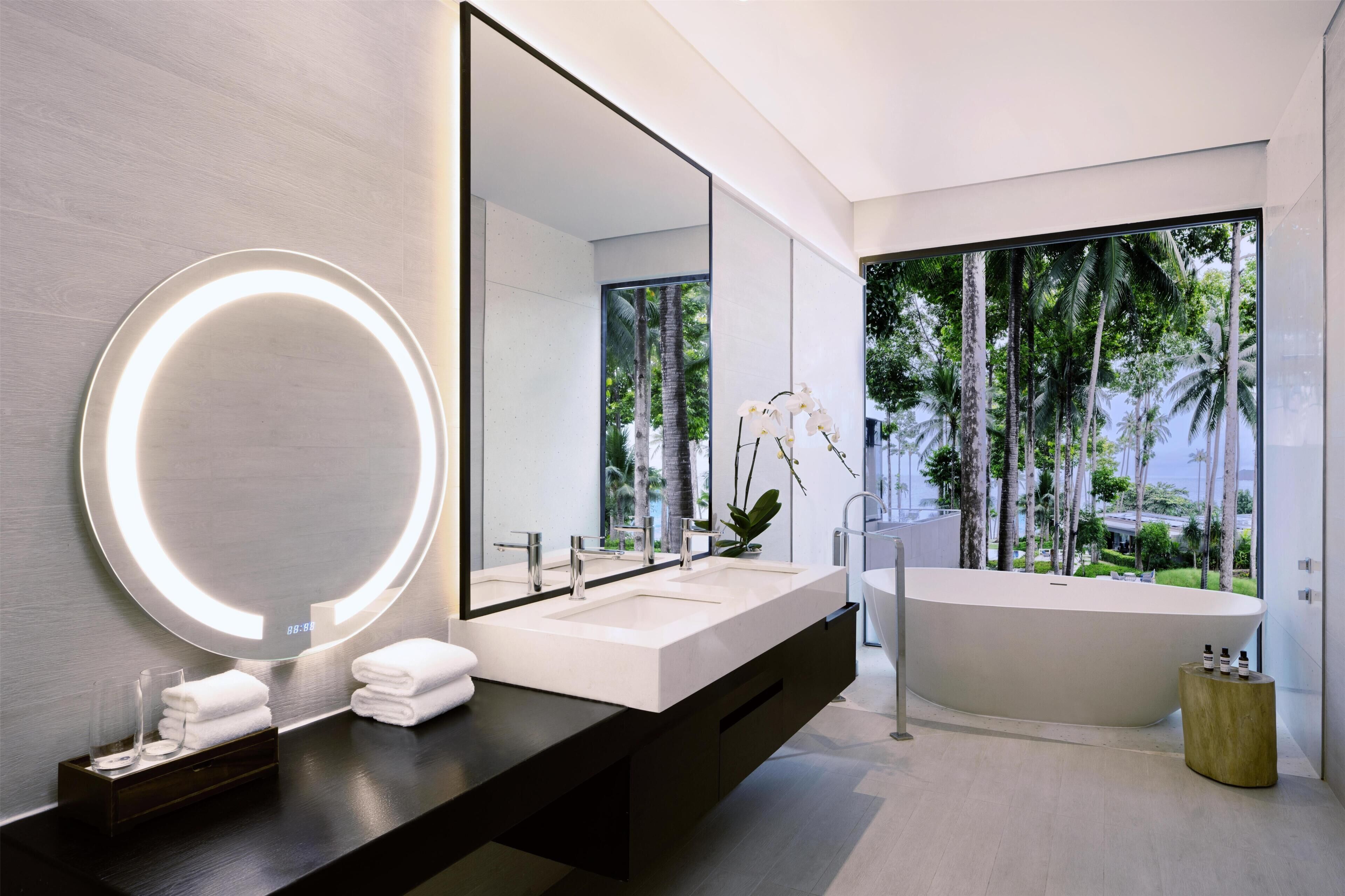 A modern bathroom with a large mirror, two sinks, a bathtub, and a view of palm trees.