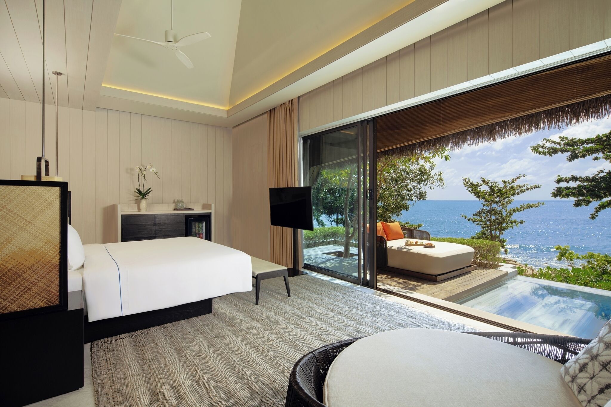 A modern hotel room with a large bed, a TV, a small table, and a view of the ocean through floor-to-ceiling windows.