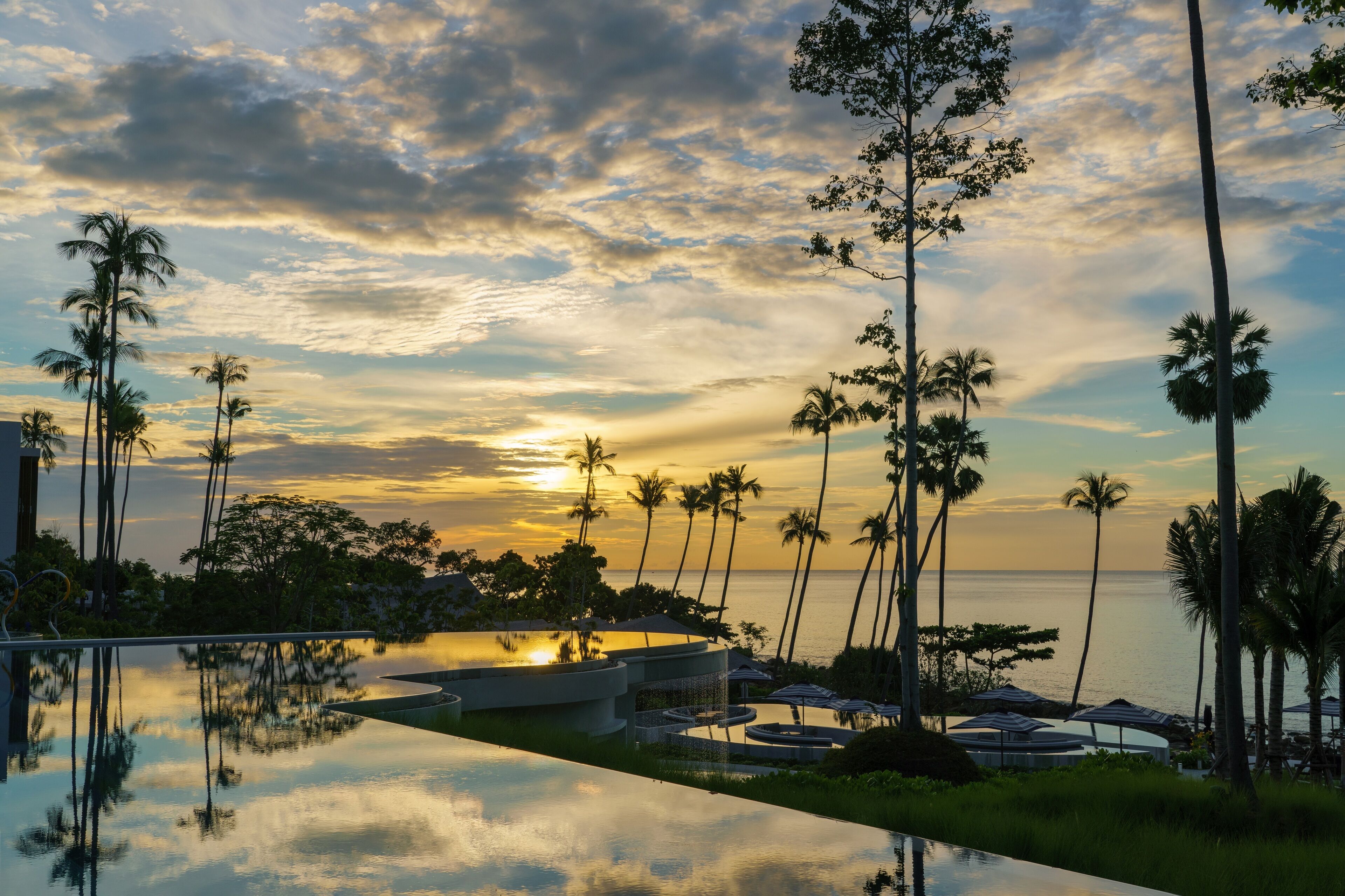A resort with a pool reflecting palm trees and a sunset over the ocean.