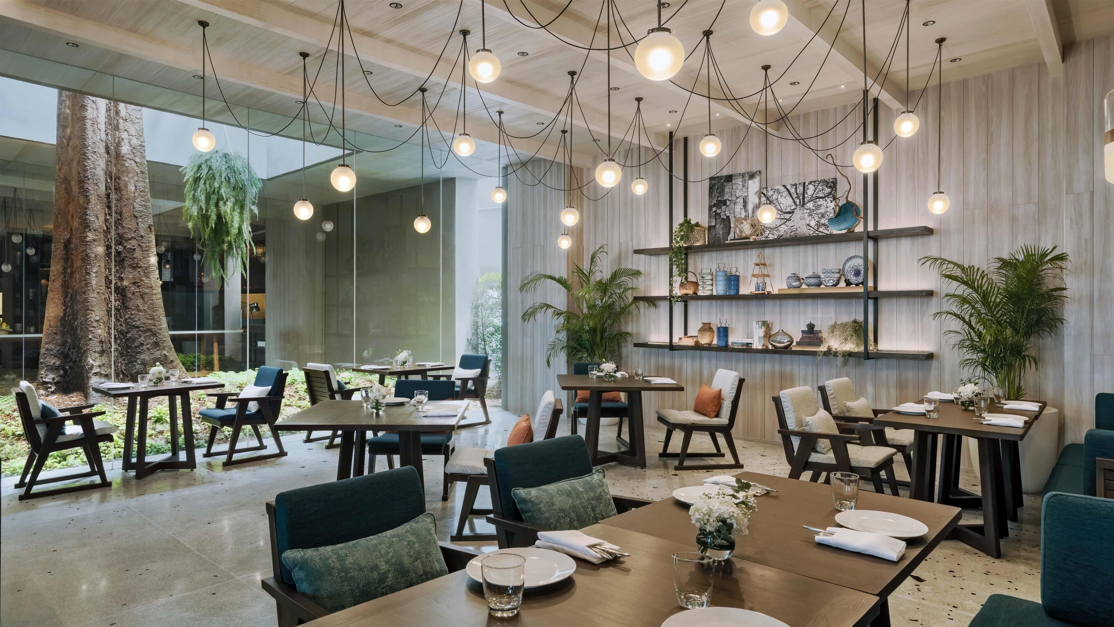 Restaurante moderno com várias mesas, cadeiras, luminárias pendentes e prateleiras com objetos decorativos.