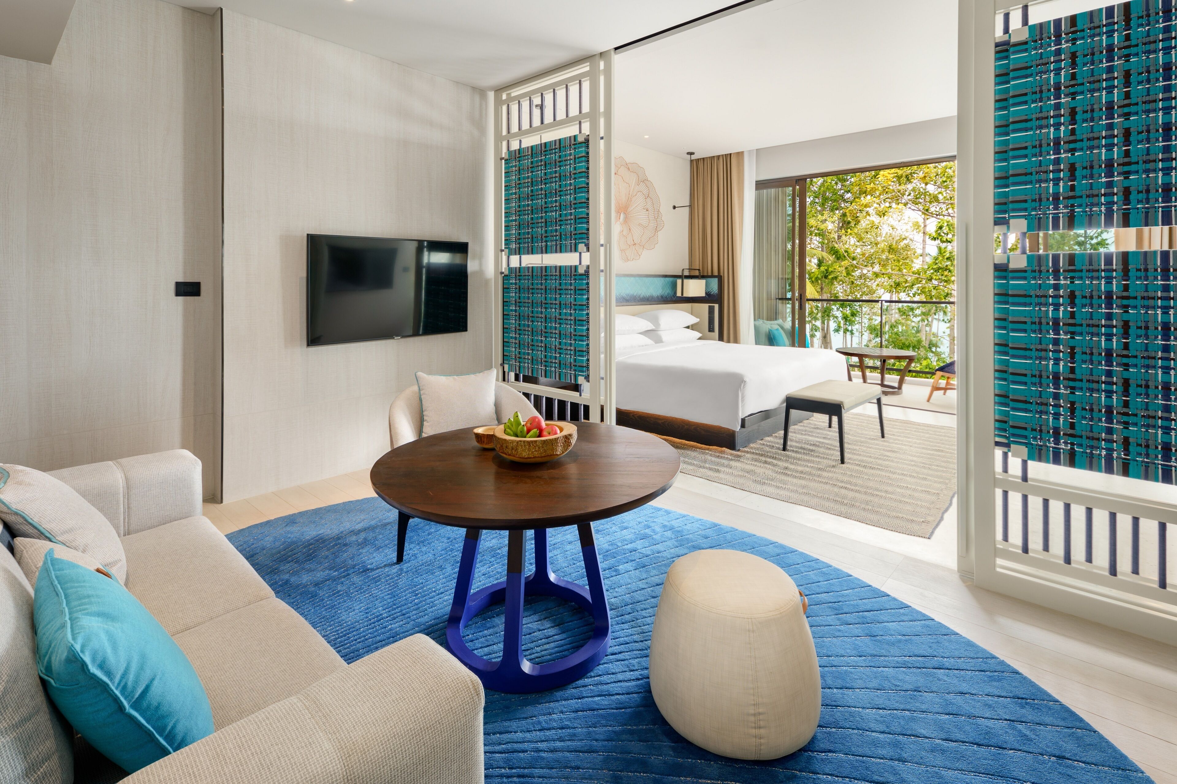 A modern hotel room with a large bed, a round table with a bowl of fruit, a sofa with cushions, and a television.