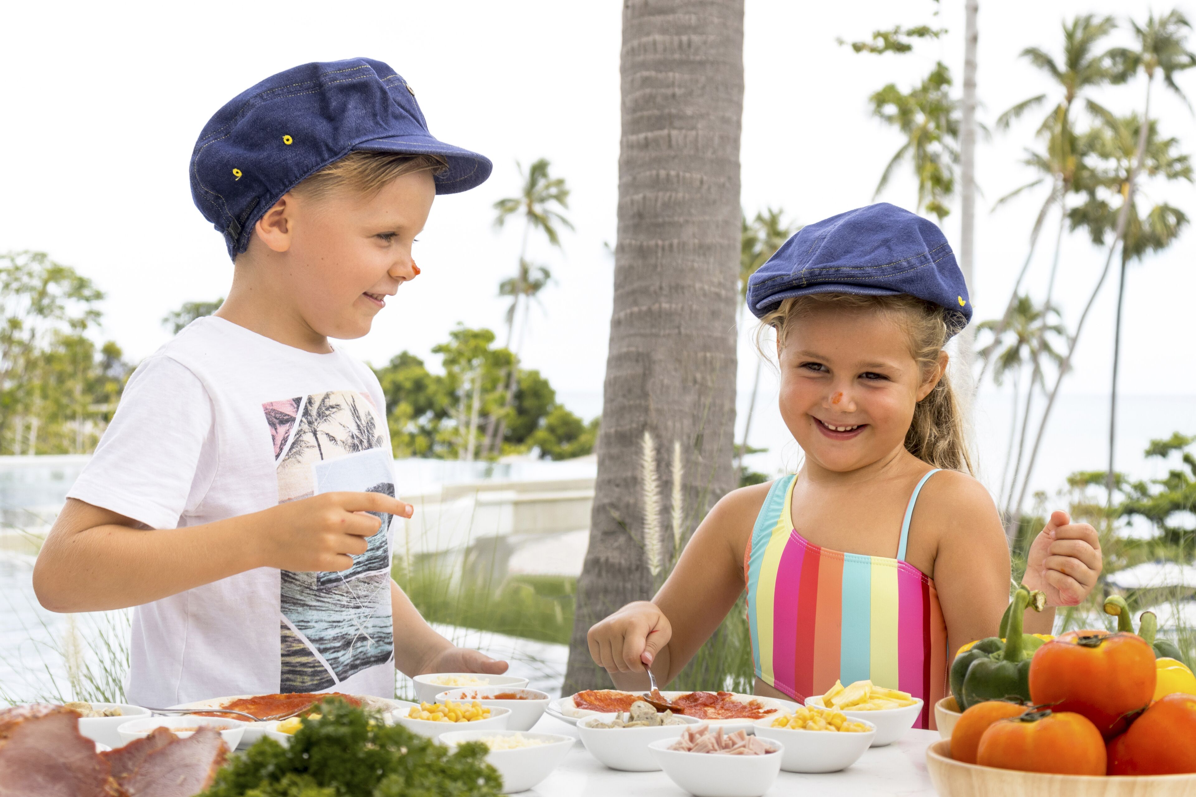Two children dining outdoors with a table full of food and a view of palm trees and the ocean.