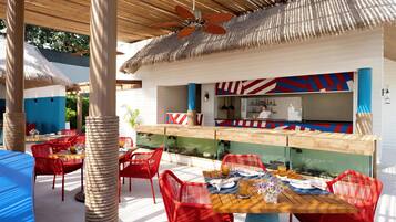 An outdoor dining area with red chairs and a wooden table, a bar area with a blue and red design, and a thatched roof structure.