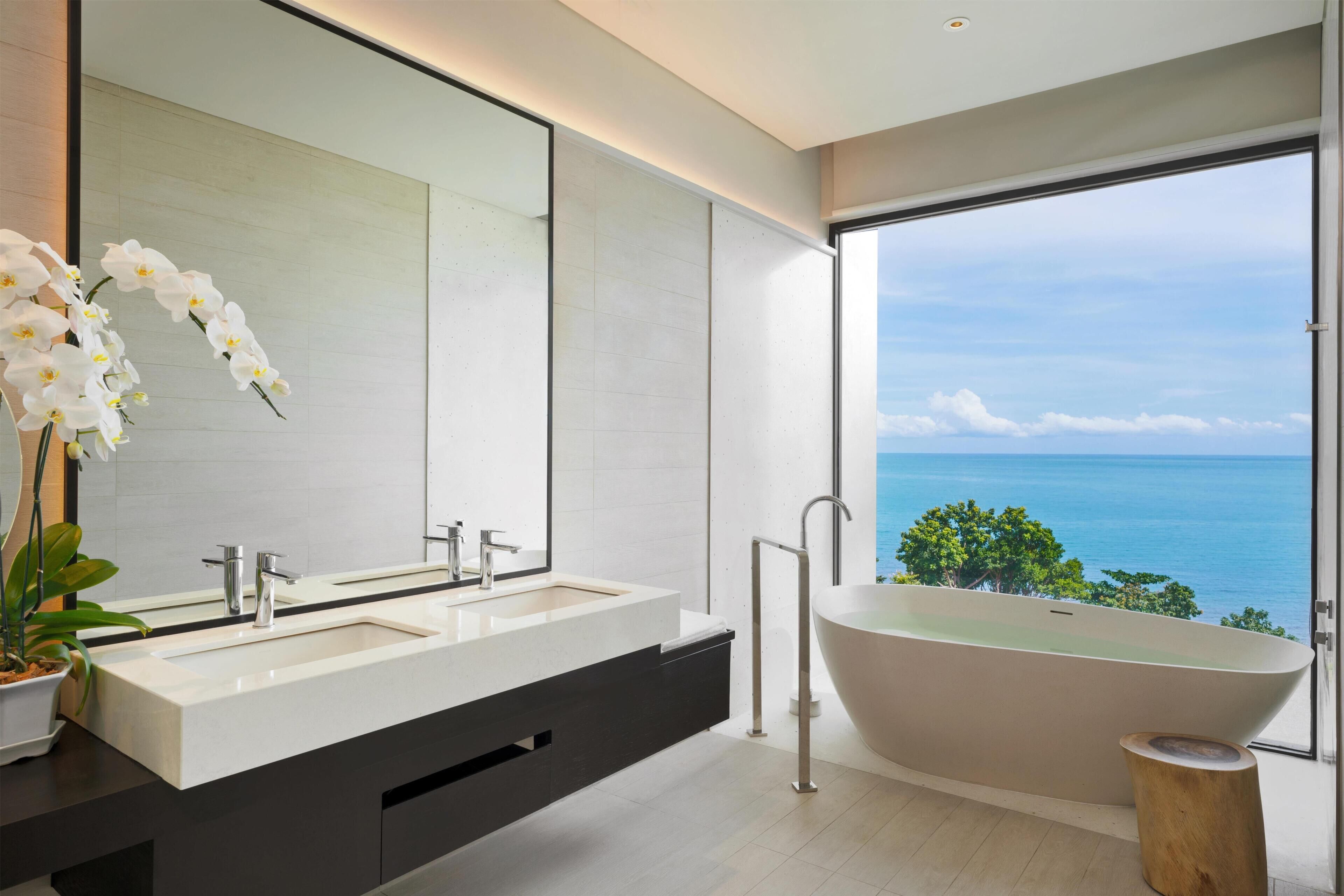 A modern bathroom with a dual sink vanity, a freestanding bathtub, and a large mirror reflecting a view of the ocean.