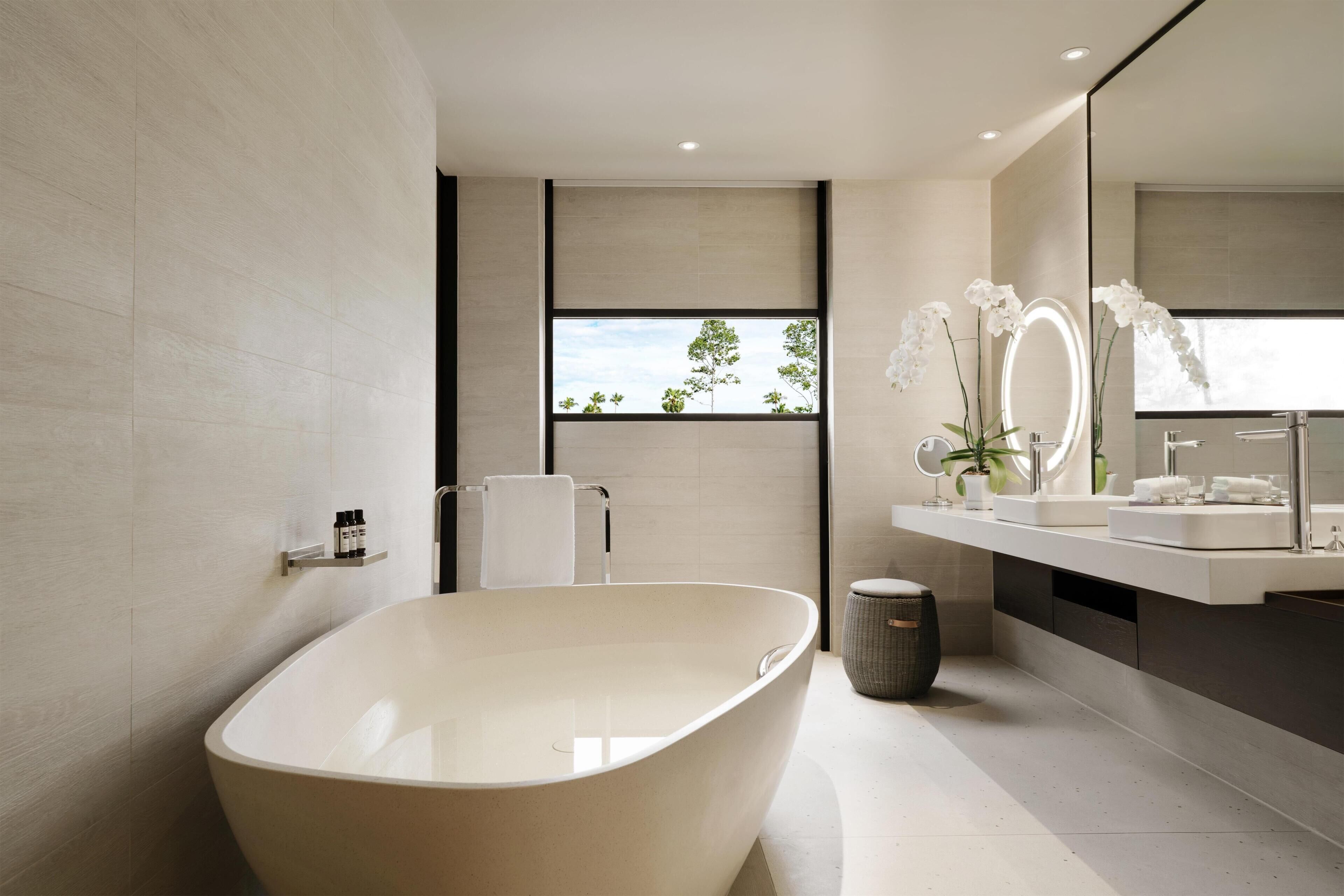 A modern bathroom with a freestanding bathtub, a large mirror, and a window with a view of trees.