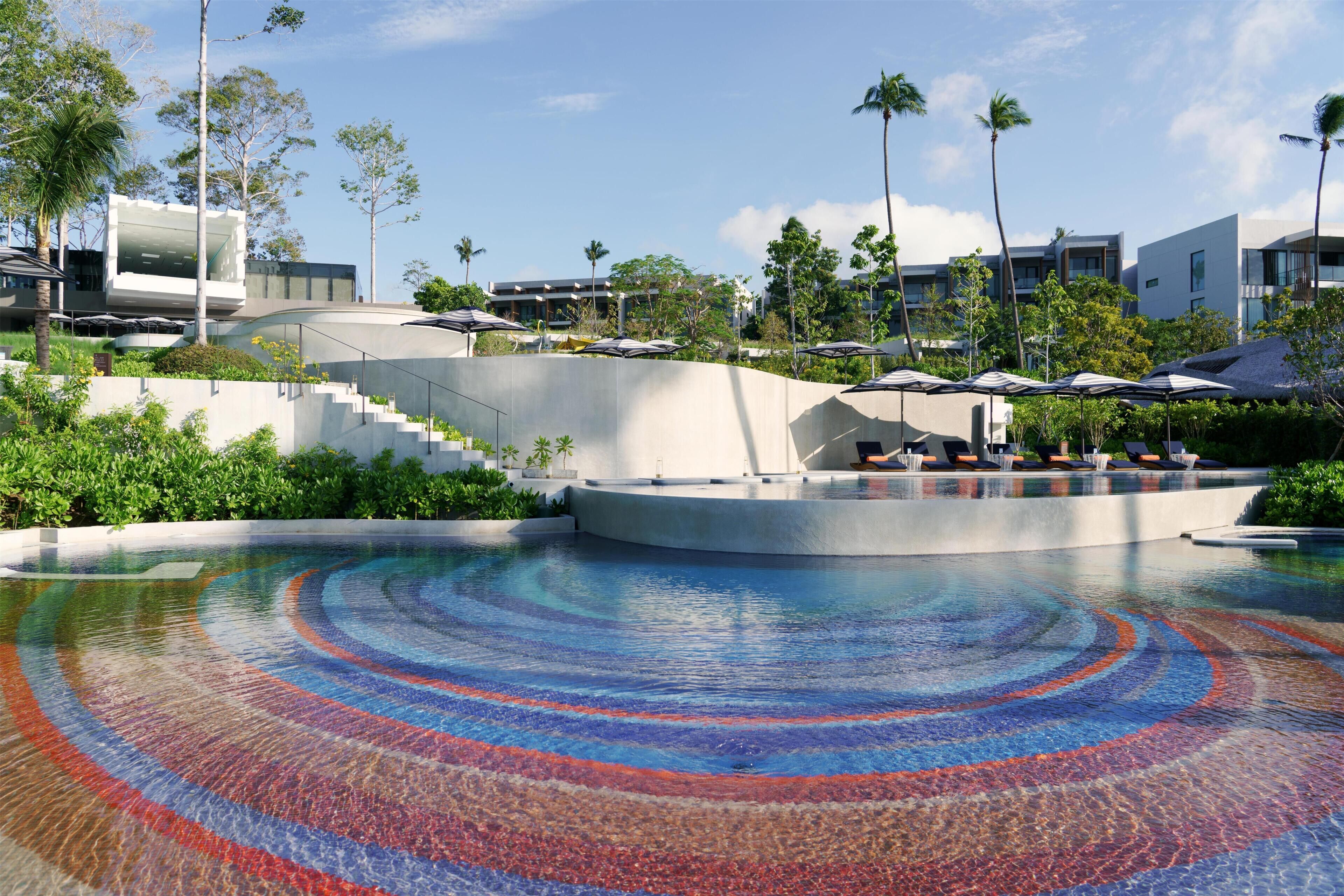 A swimming pool with a colorful mosaic design, surrounded by a landscaped area with palm trees and modern buildings.