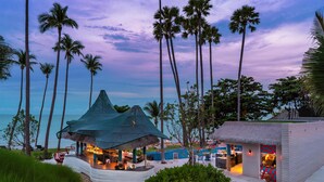 Un restaurante frente al mar con techo de paja, palmeras y un área de piscina.