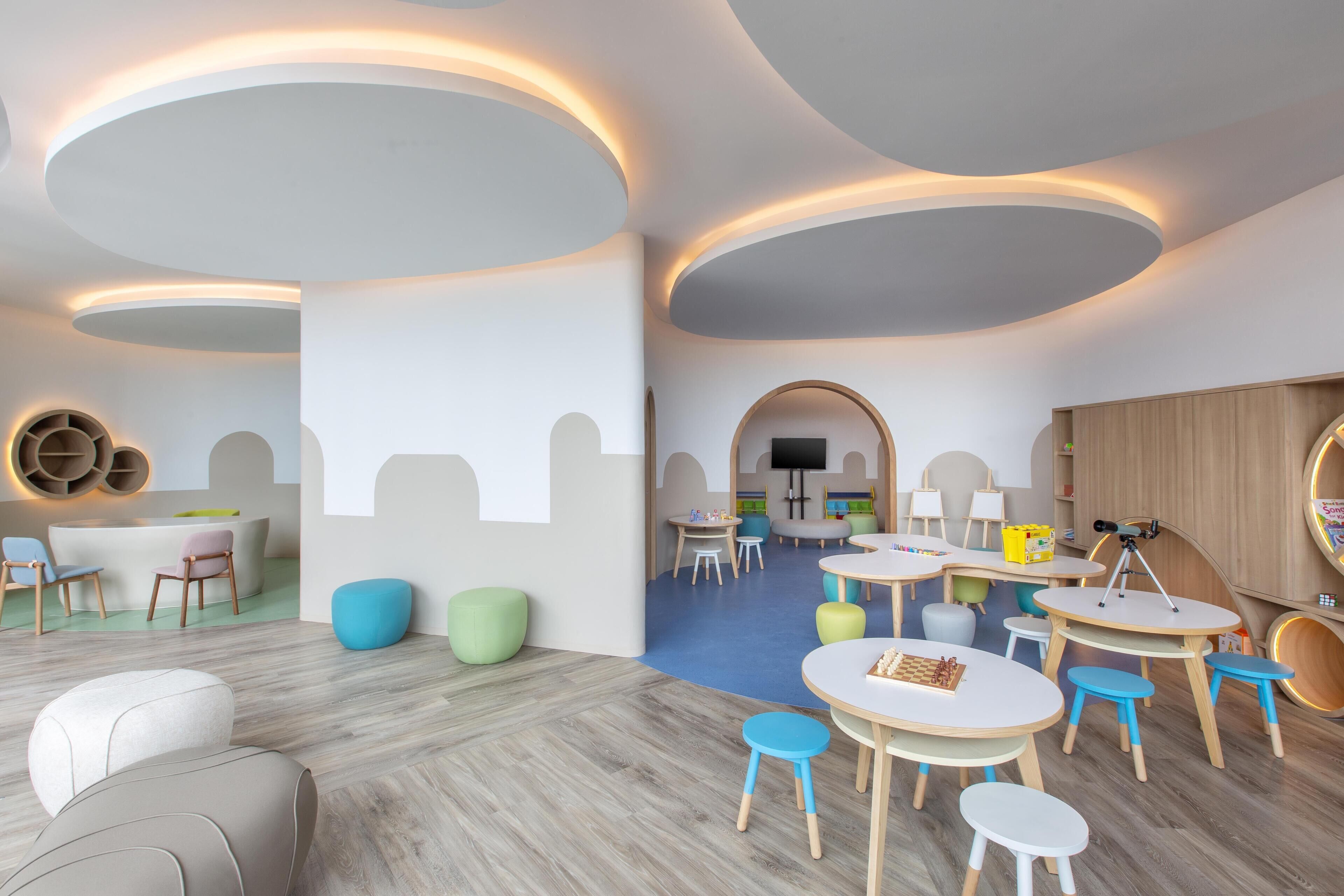 A children's playroom with round tables, colorful chairs, and a TV in the background.