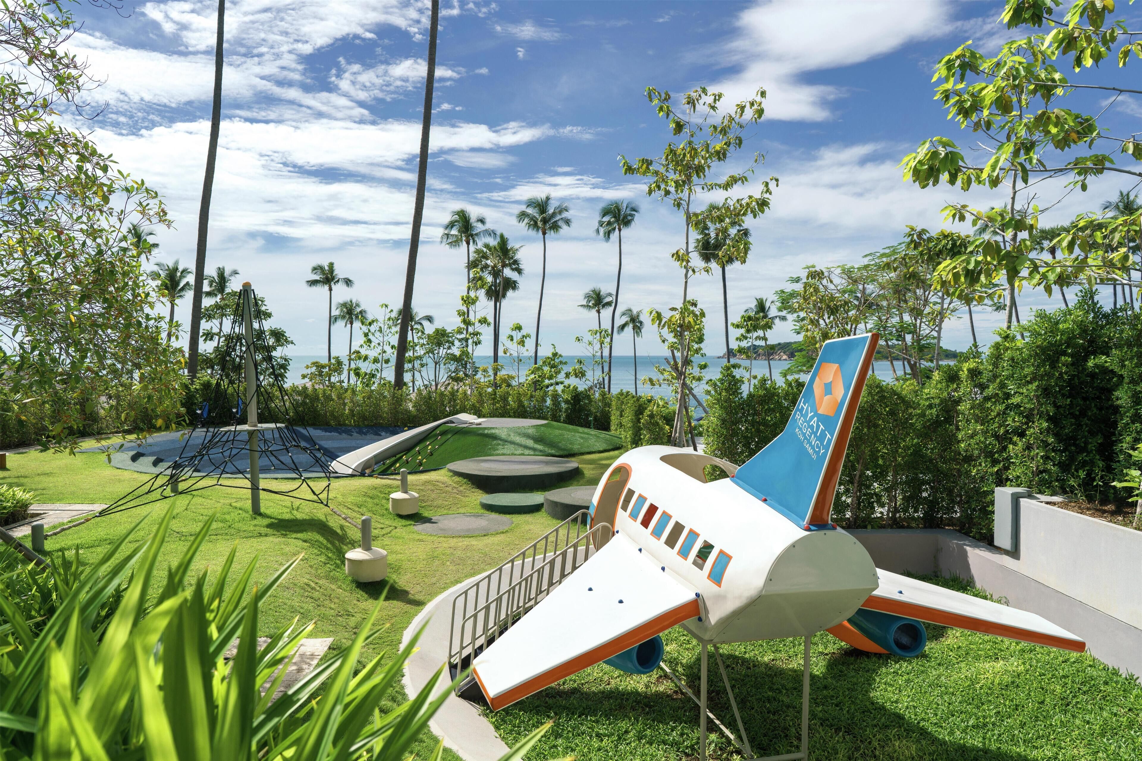 A playground with a slide and climbing structures, surrounded by palm trees and a view of the ocean.