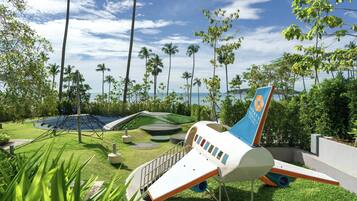 A playground with a slide and climbing structures, surrounded by palm trees and a view of the ocean.