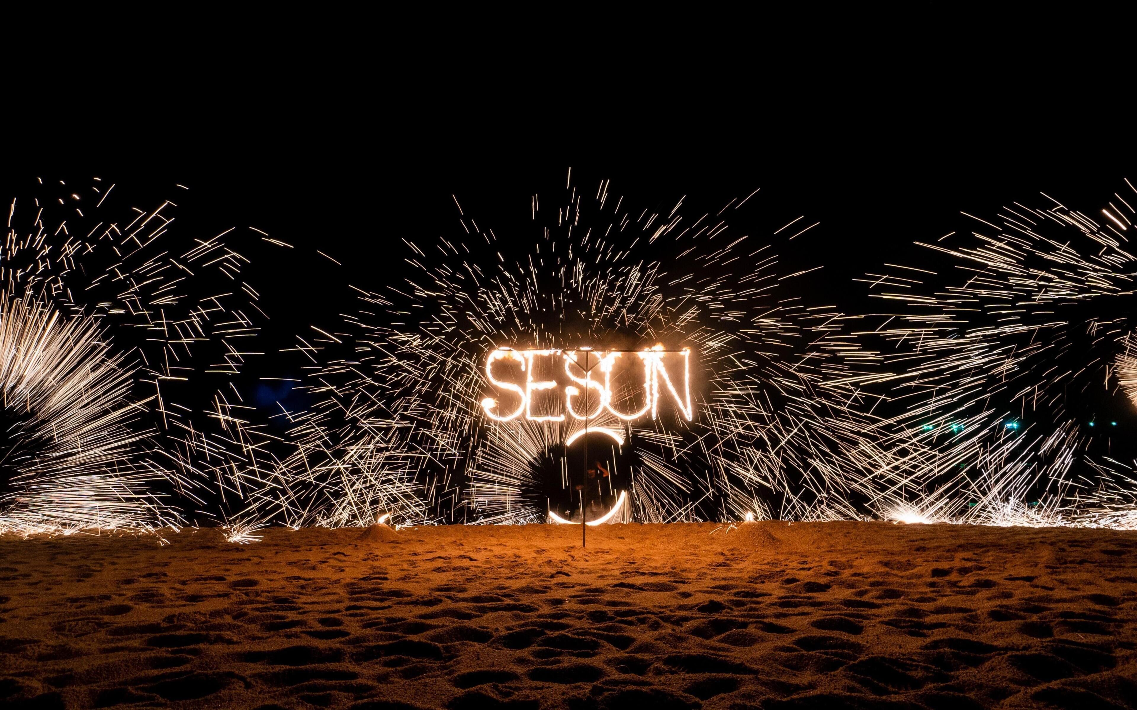 A nighttime beach scene with fireworks and the word 'SEASON' illuminated in the sand.