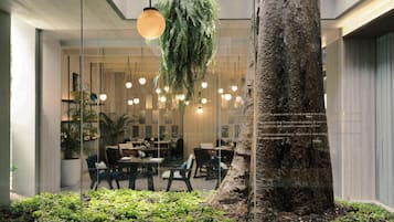 Un espacio interior moderno con un gran árbol, plantas colgantes y una zona de comedor con mesas y sillas.