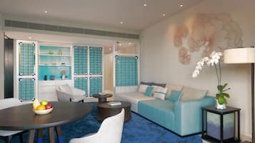 A modern living room with a blue carpet, a round dining table with fruit, a sofa, and a decorative wall piece.