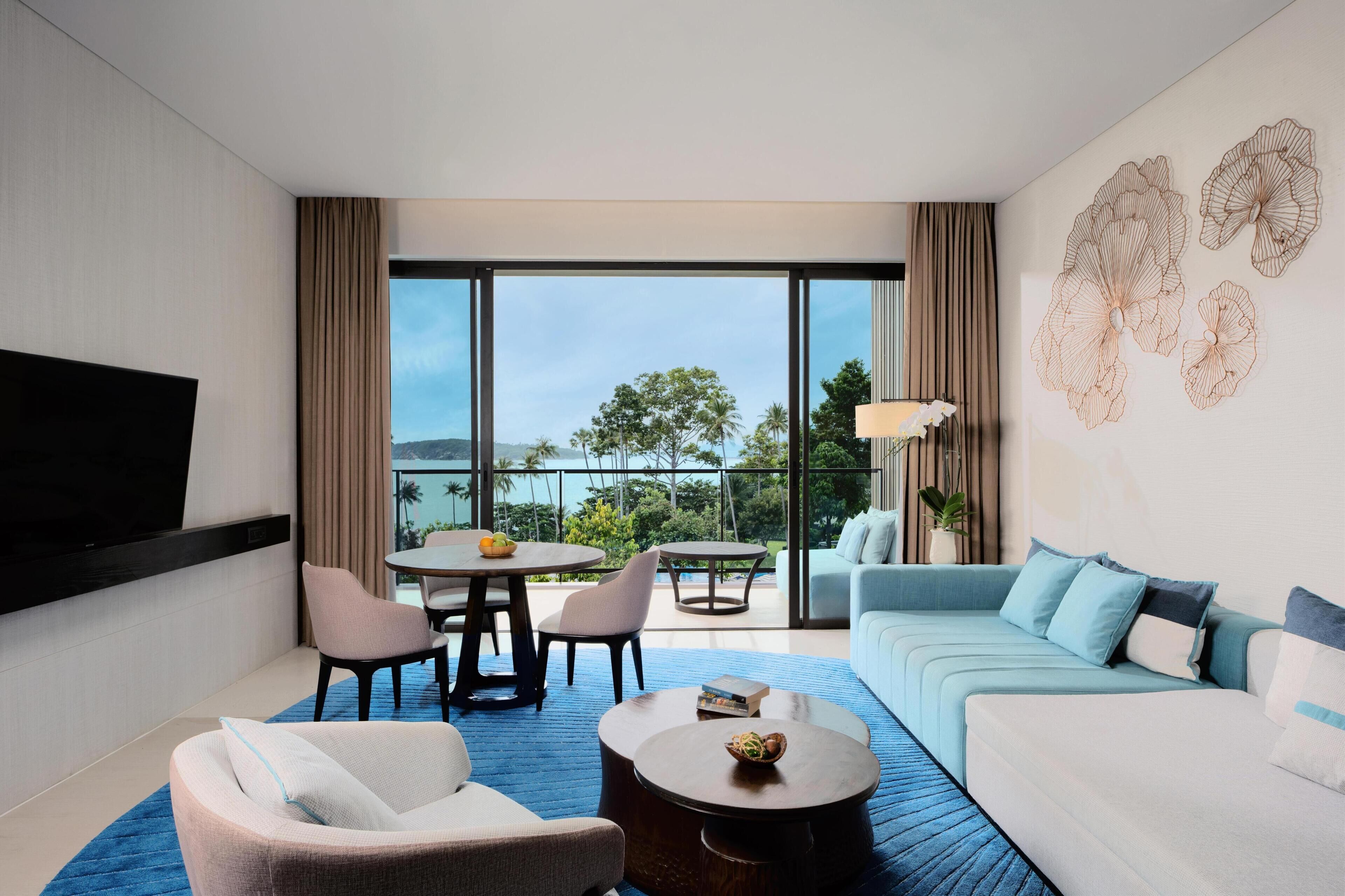 Una habitación de hotel moderna con televisión de pantalla plana, un sofá, una mesa de comedor con sillas y un balcón con vista al mar.